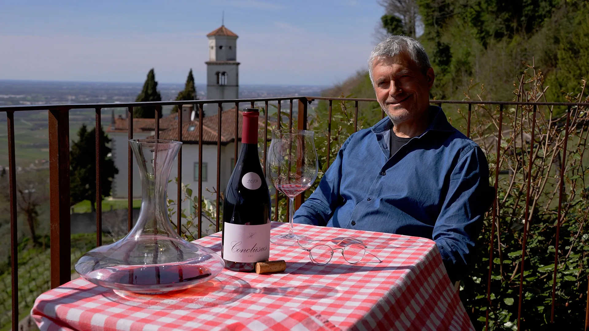 Man in blue shirt smiling at an outdoor table with a bottle of red wine, glasses, and a carafe on a red‑and‑white checkered tablecloth, hillside view behind a railing.