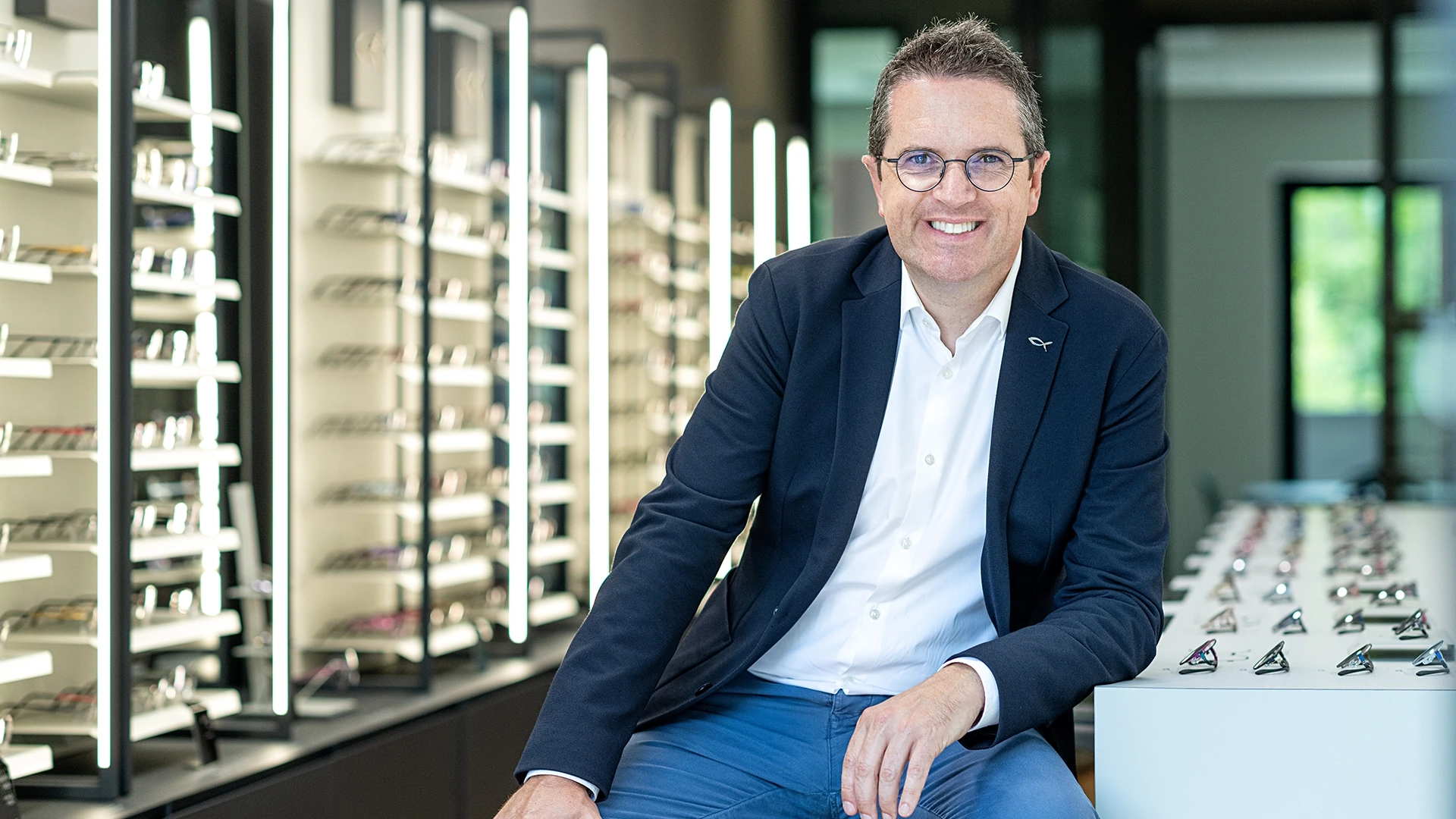 Smiling man in a blazer and white shirt sits at a white display table with rows of eyeglasses behind him in a modern eyewear store.