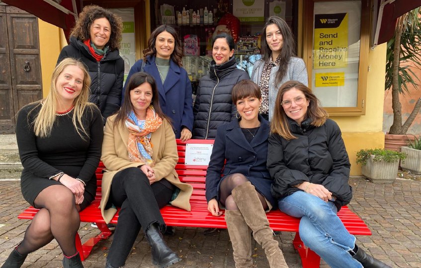 Eight women pose together on a bright red bench outside a shop, smiling at the camera.