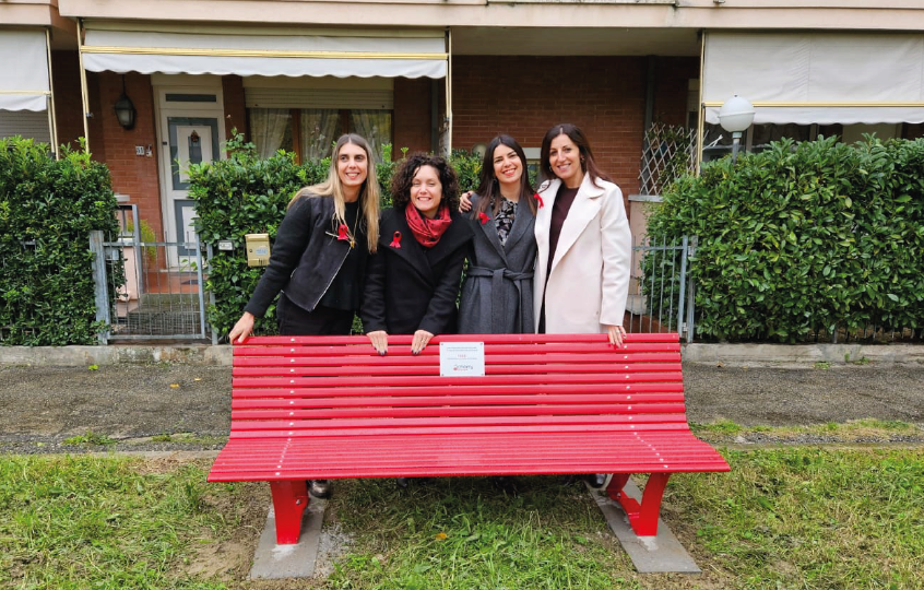 Four women stand behind a bright red bench on a sidewalk, smiling at the camera in a residential area with hedges.