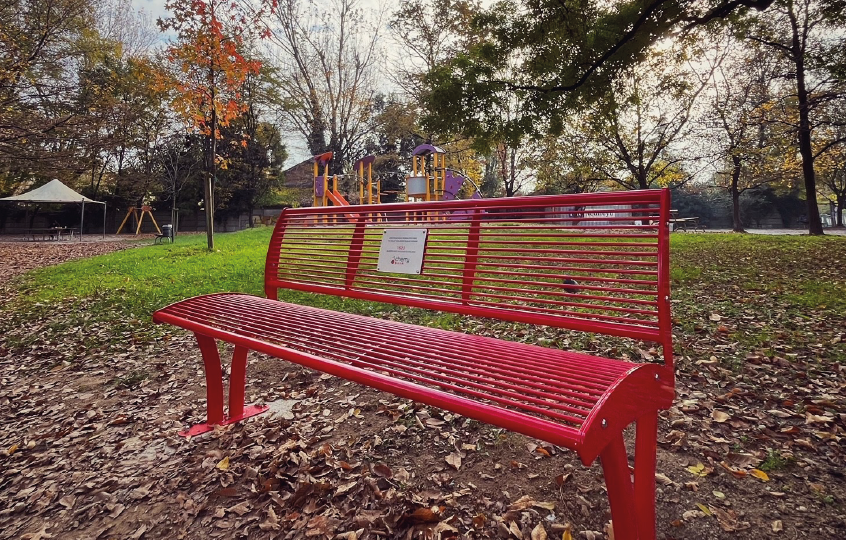 Red metal park bench on a leaf-covered ground with a playground in the background during autumn