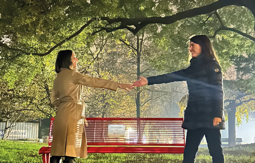 Two women greet with a handshake beside a red park bench amid sunlit trees.