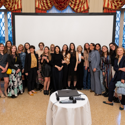 Group of about 25 women posing for a photo in a room with a blank projector screen behind them; a table with a microphone and laptop is in the foreground.