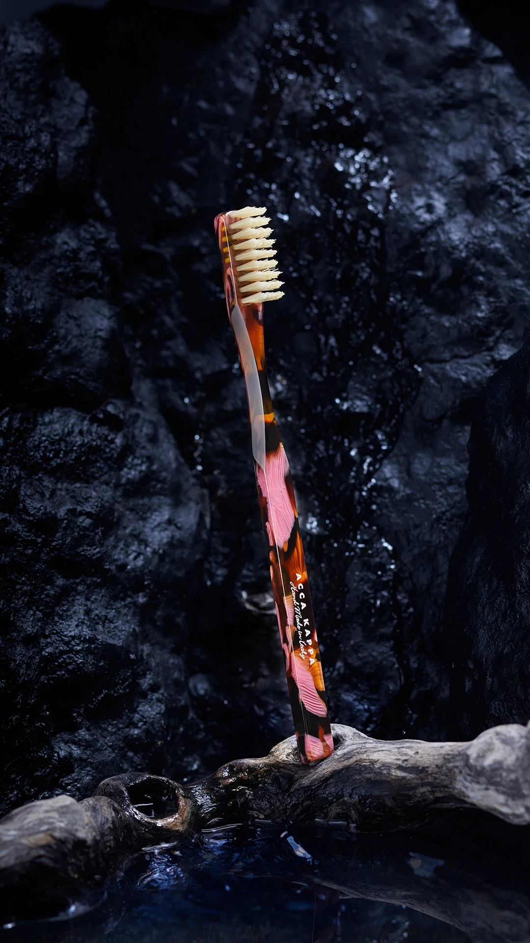 Colorful marbled toothbrush with beige bristles standing upright against a dark rocky background near water.
