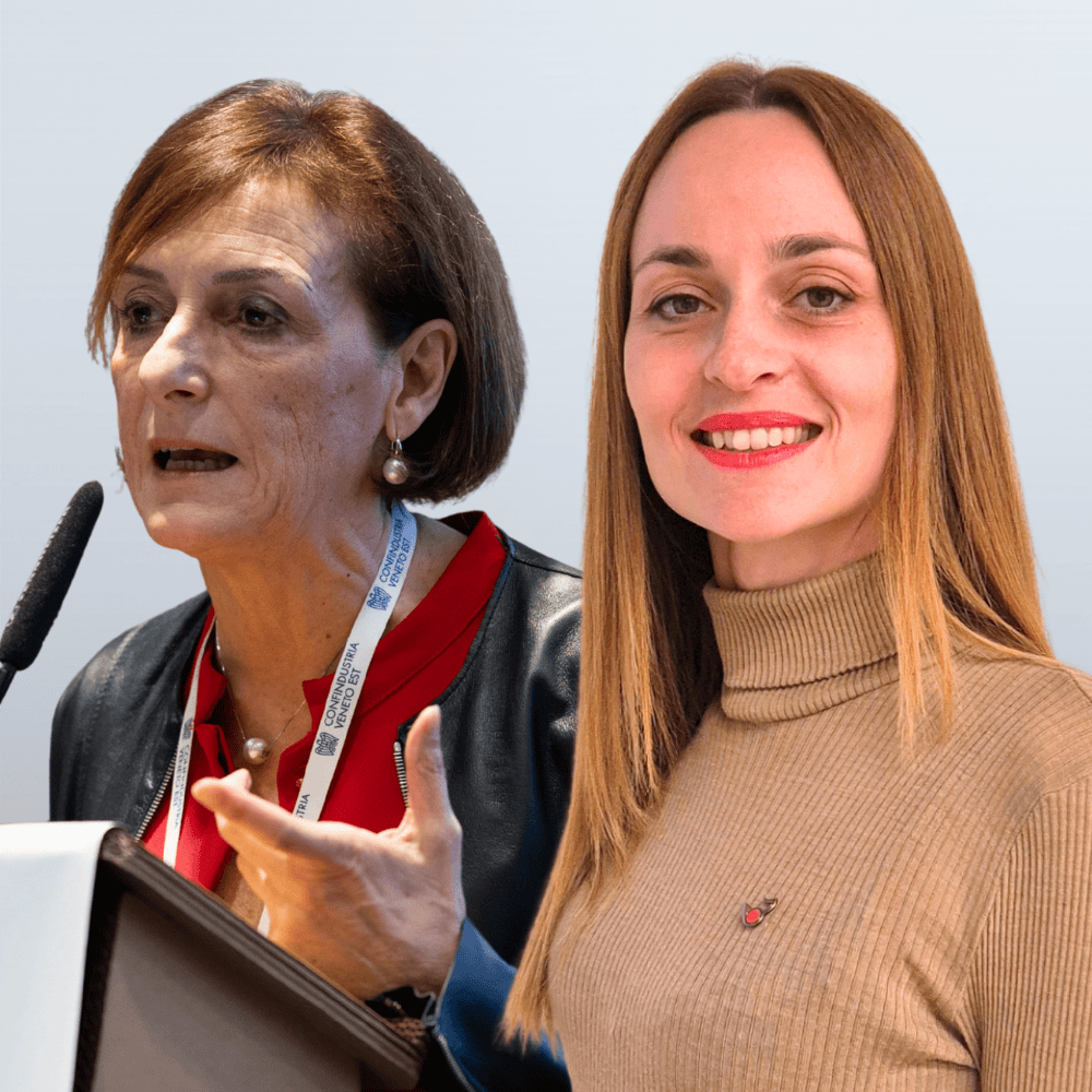Two women at a podium during a conference; one speaks into a microphone while the other smiles at the camera.