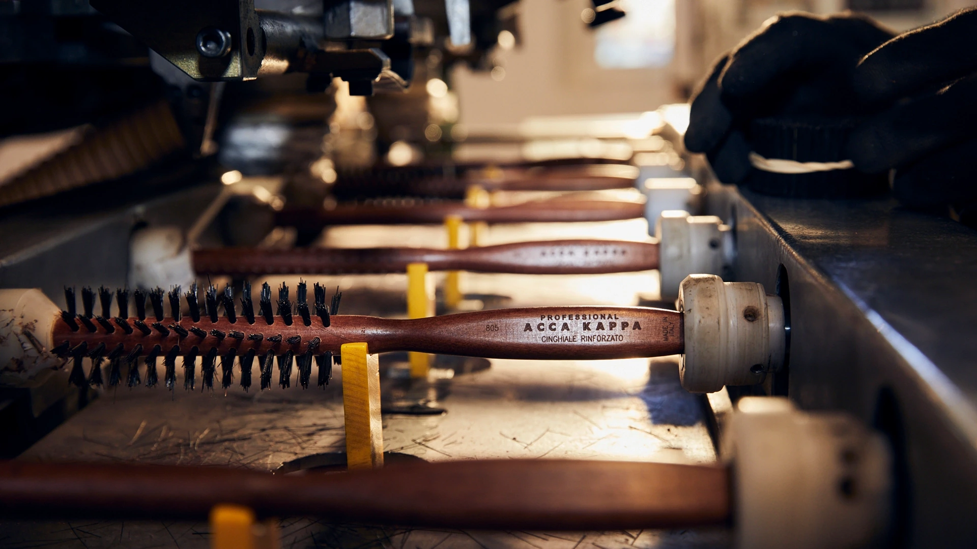 Row of wooden-handled brushes on a production line in a workshop setting, with bristles exposed and factory equipment nearby.