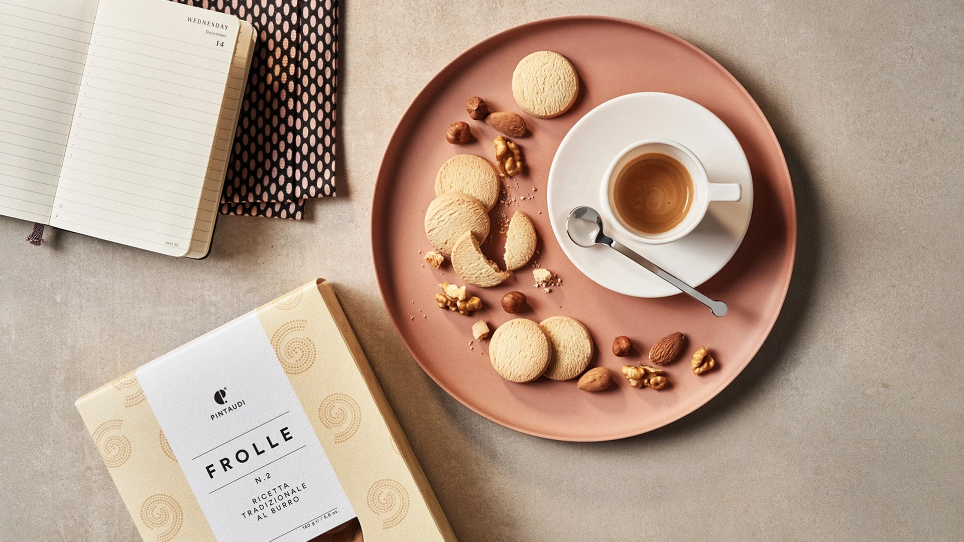 A flat lay of a box of cookies beside a notebook and a pink plate with cookies and an espresso cup with a spoon.