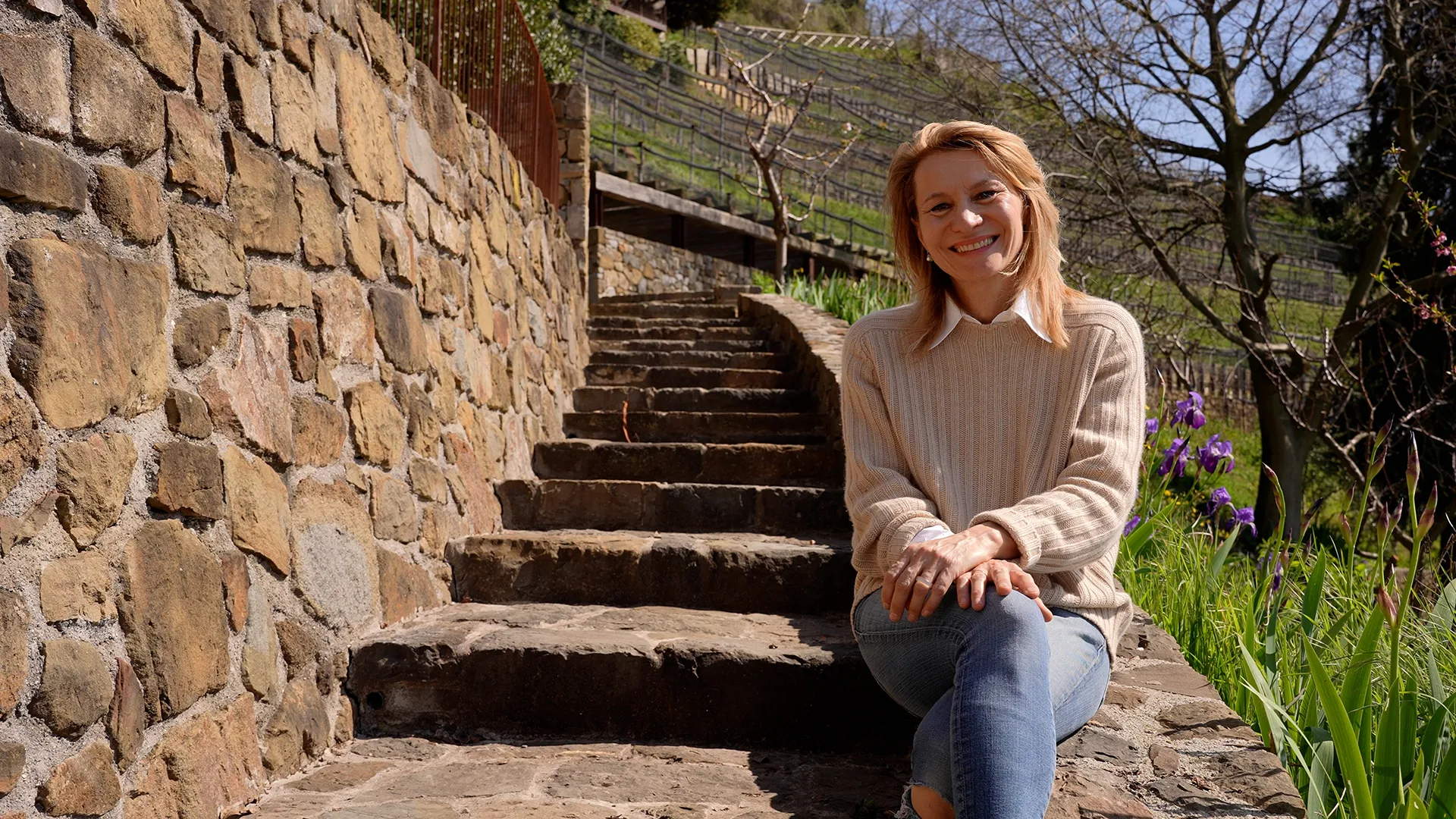 Smiling woman seated on stone steps beside a rough stone wall in a sunny garden with purple irises nearby.