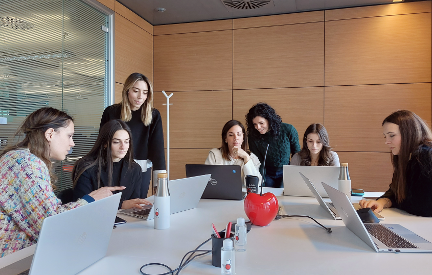 Seven women collaborating around a white conference table with laptops in a modern office, one standing.By the wall with wooden panels, a diverse team discusses a project.