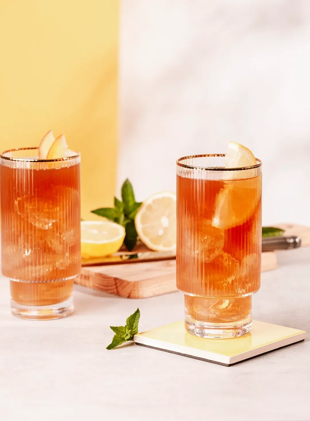 Two pink drinks in short glasses with lemon wedges and mint, set on a light table with a wooden board nearby.
