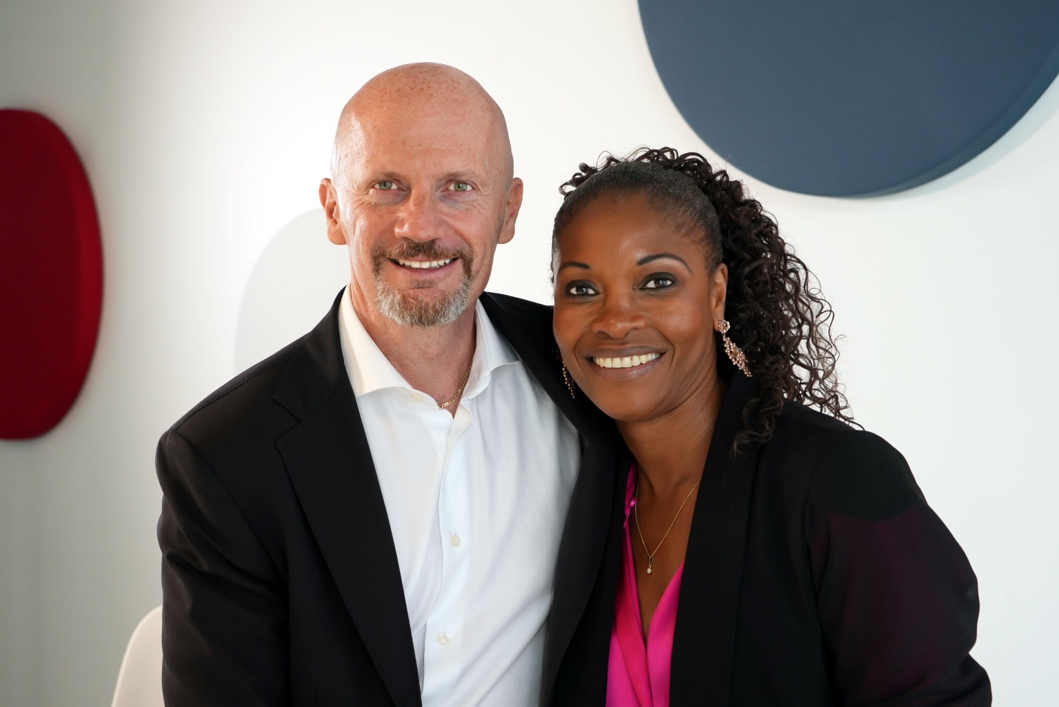 Two smiling colleagues posing together in business attire, leaning close in a bright, modern setting with circular wall decor behind them.