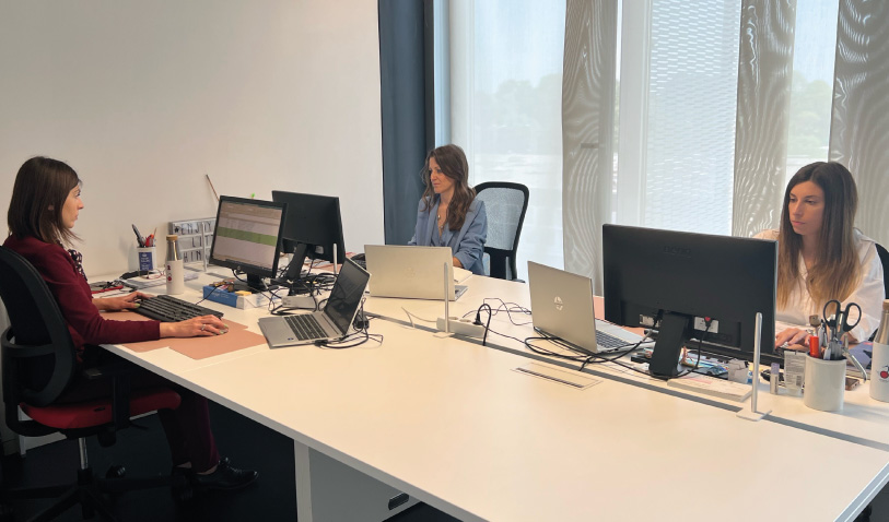 Three women working at a long white desk with laptops and monitors in a bright, modern office. One on the left types at her computer while two others sit at the center and right, facing their screens or models.