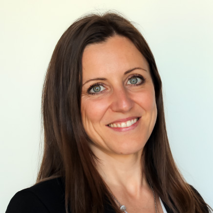 Portrait of a smiling woman with long brown hair and blue eyes, wearing a black blazer against a light neutral background.