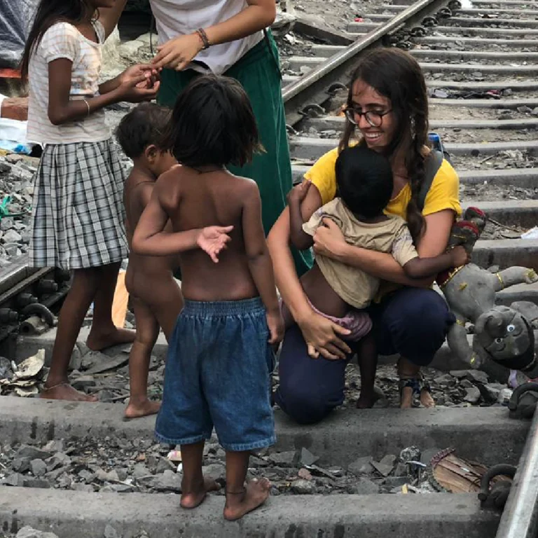 A young woman wearing glasses and a yellow shirt crouches on railroad tracks, cradling a small child while several barefoot children stand nearby in dusty rubble.