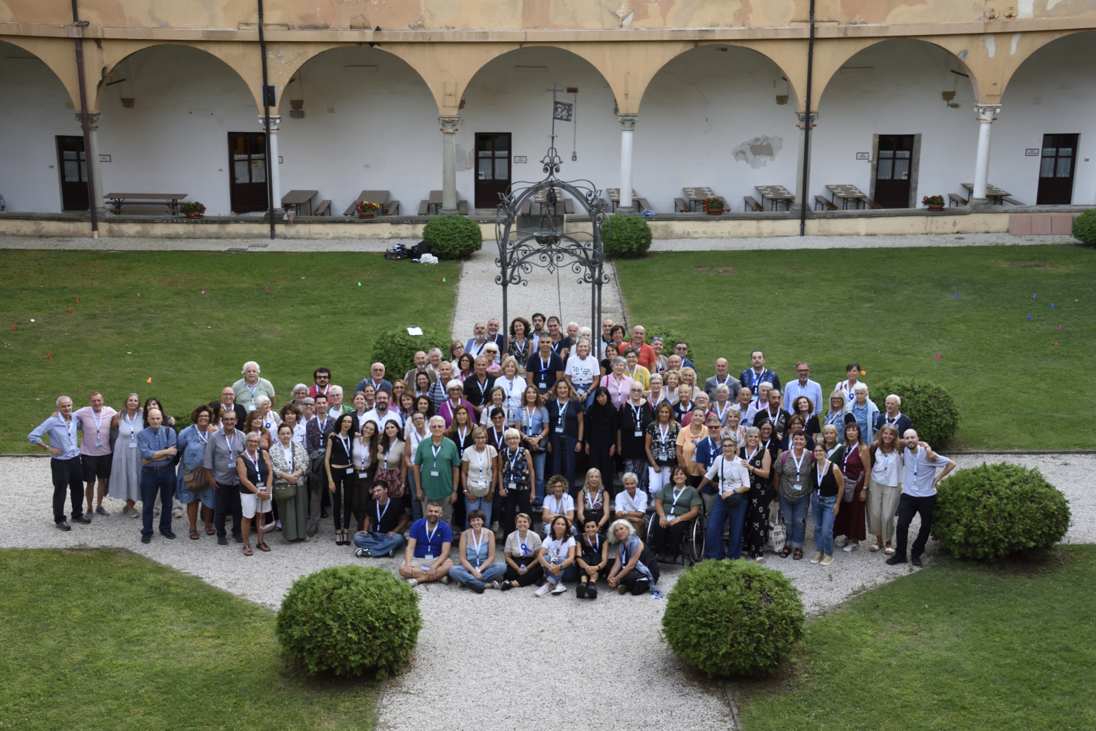 Large group of people posing for a group photo in a courtyard with arches, gravel path, and trimmed bushes; many wear name badges.