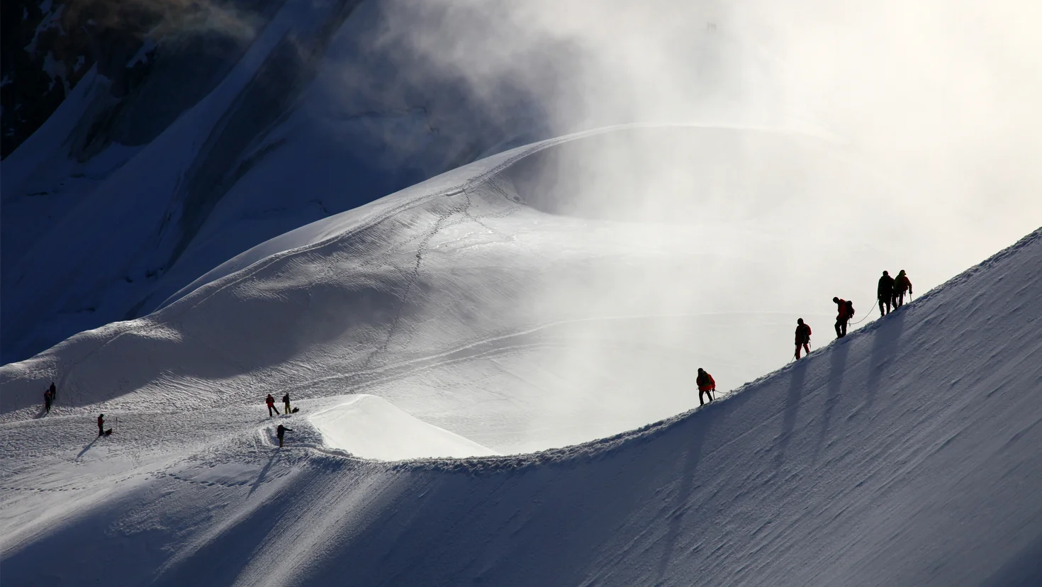 Group of climbers ascending a snow-covered mountain ridge, footprints trailing behind them against a misty, sunlit slope.