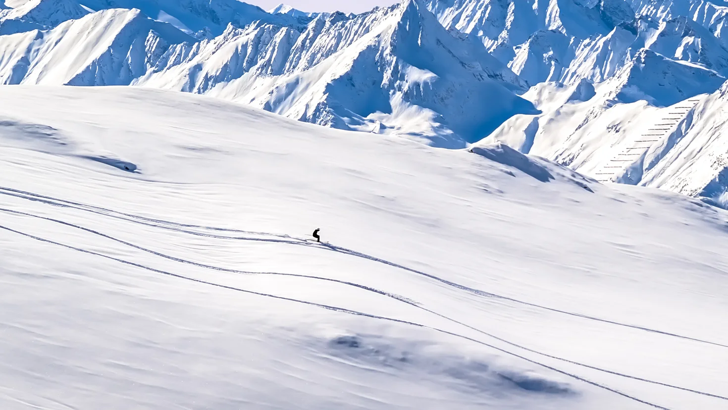 A lone skier carving a line across a snow-covered mountain slope with jagged blue peaks in the background.