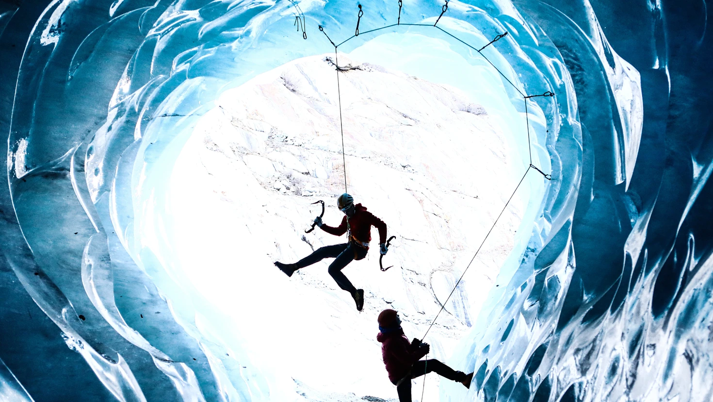 Two climbers tethered in a glowing blue ice cave; one hangs mid‑air while the other manages the rope below.