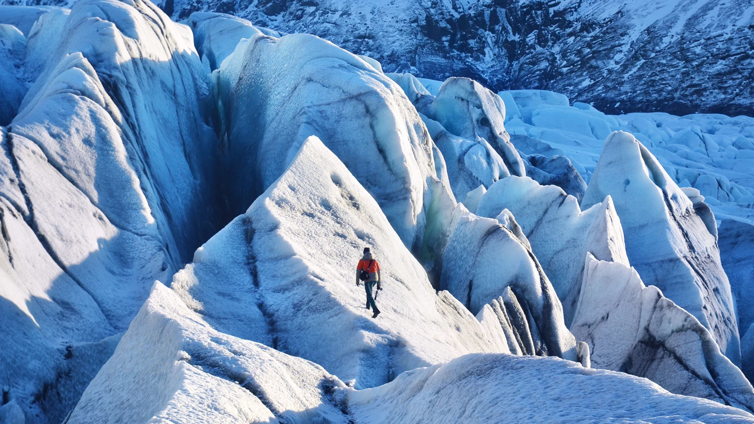 Hiker in an orange jacket walking across jagged blue glacier ice