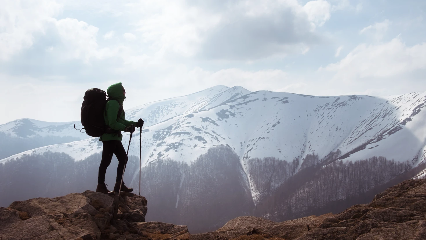 Hiker in a green jacket with a large backpack standing on a rocky cliff, gazing at snow-covered mountains under a blue sky.