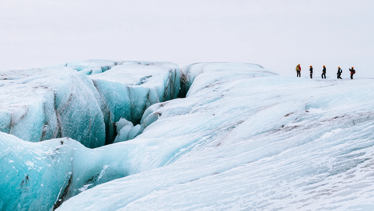 A line of climbers in bright gear walk along a vast blue glacier with deep crevasses to the left