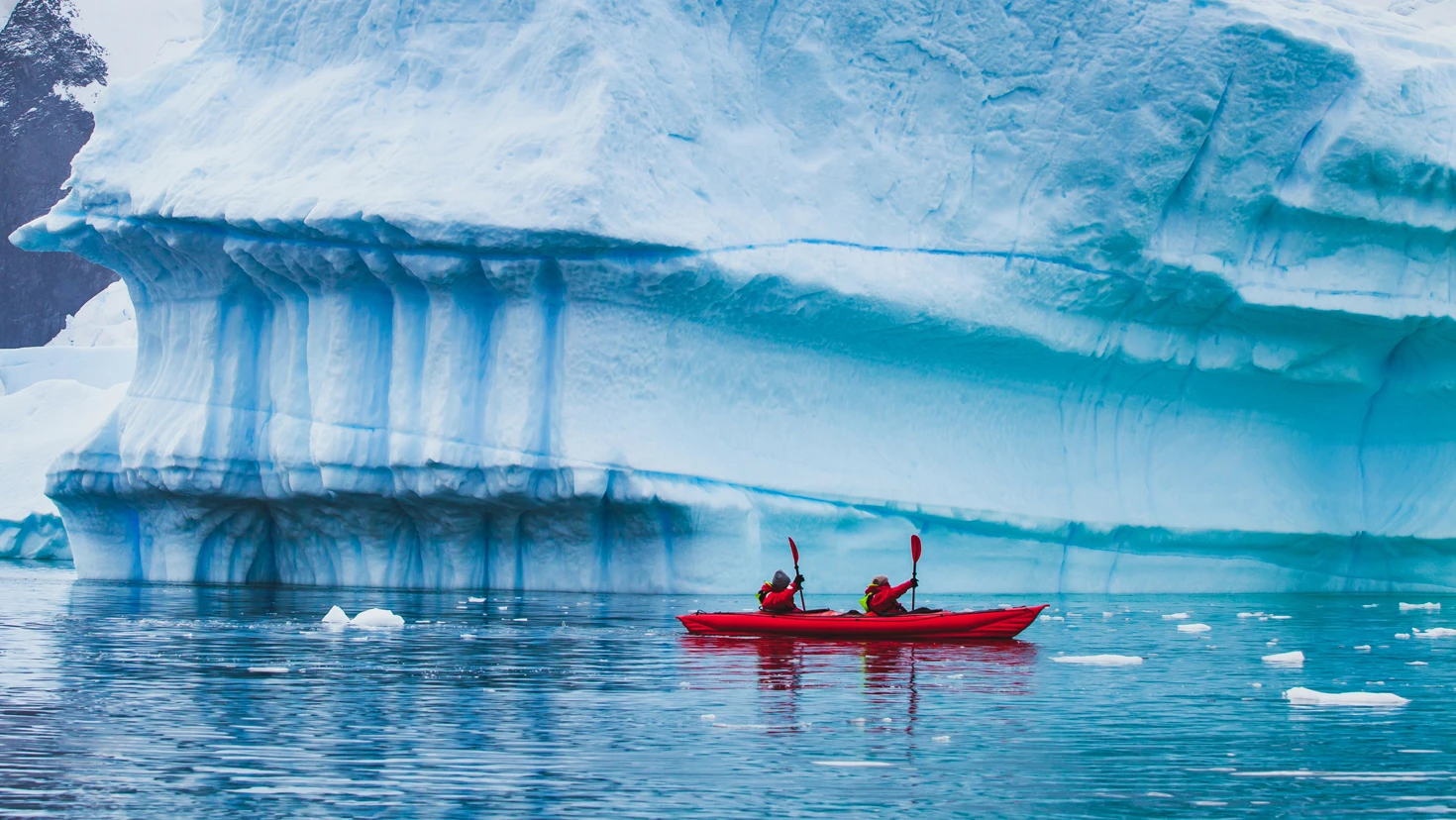 Two people in a red tandem kayak paddling near a towering blue iceberg in calm Arctic waters.