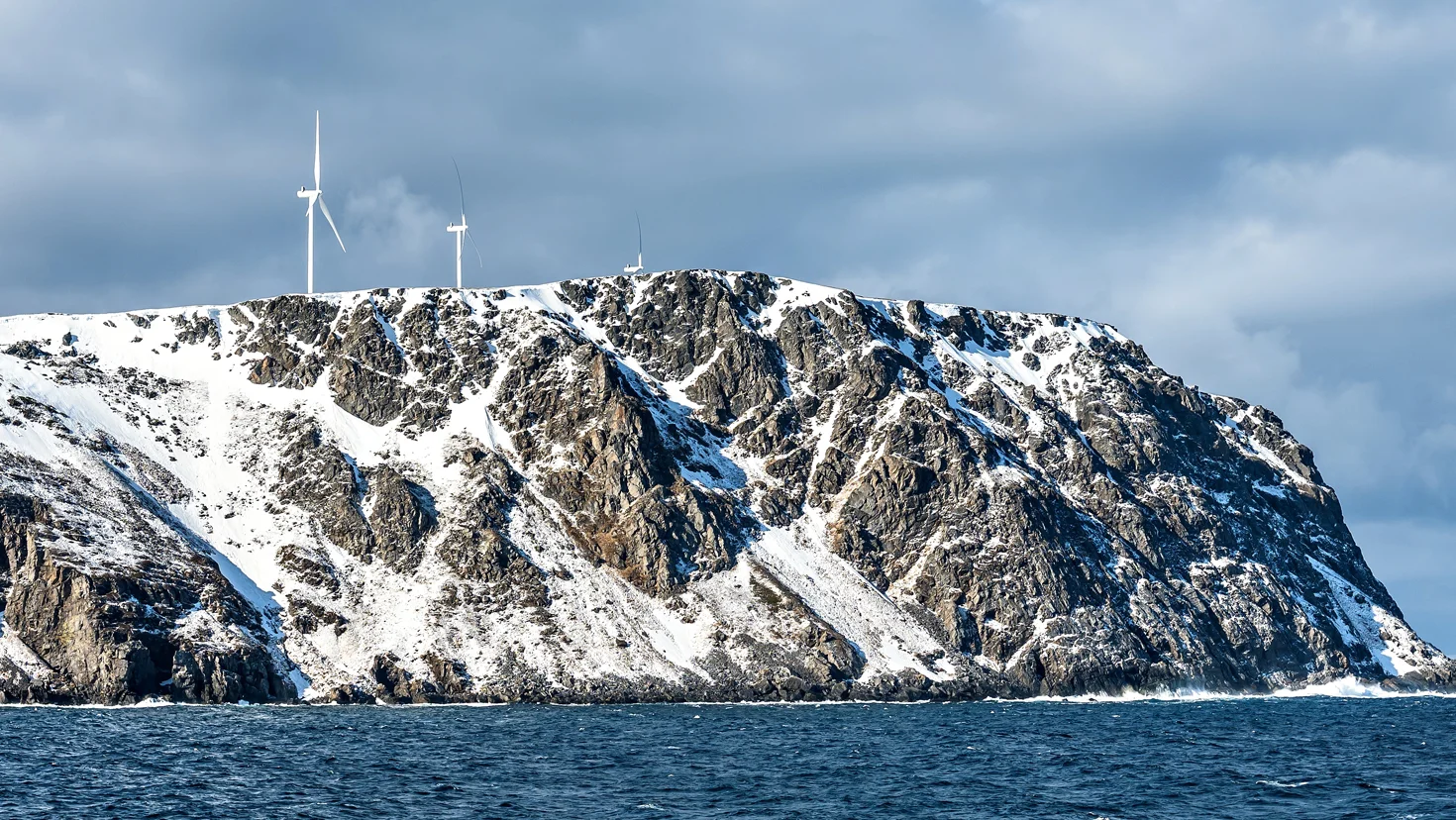 Snowy rocky island with three wind turbines on a cliff above the sea under a cloudy sky.